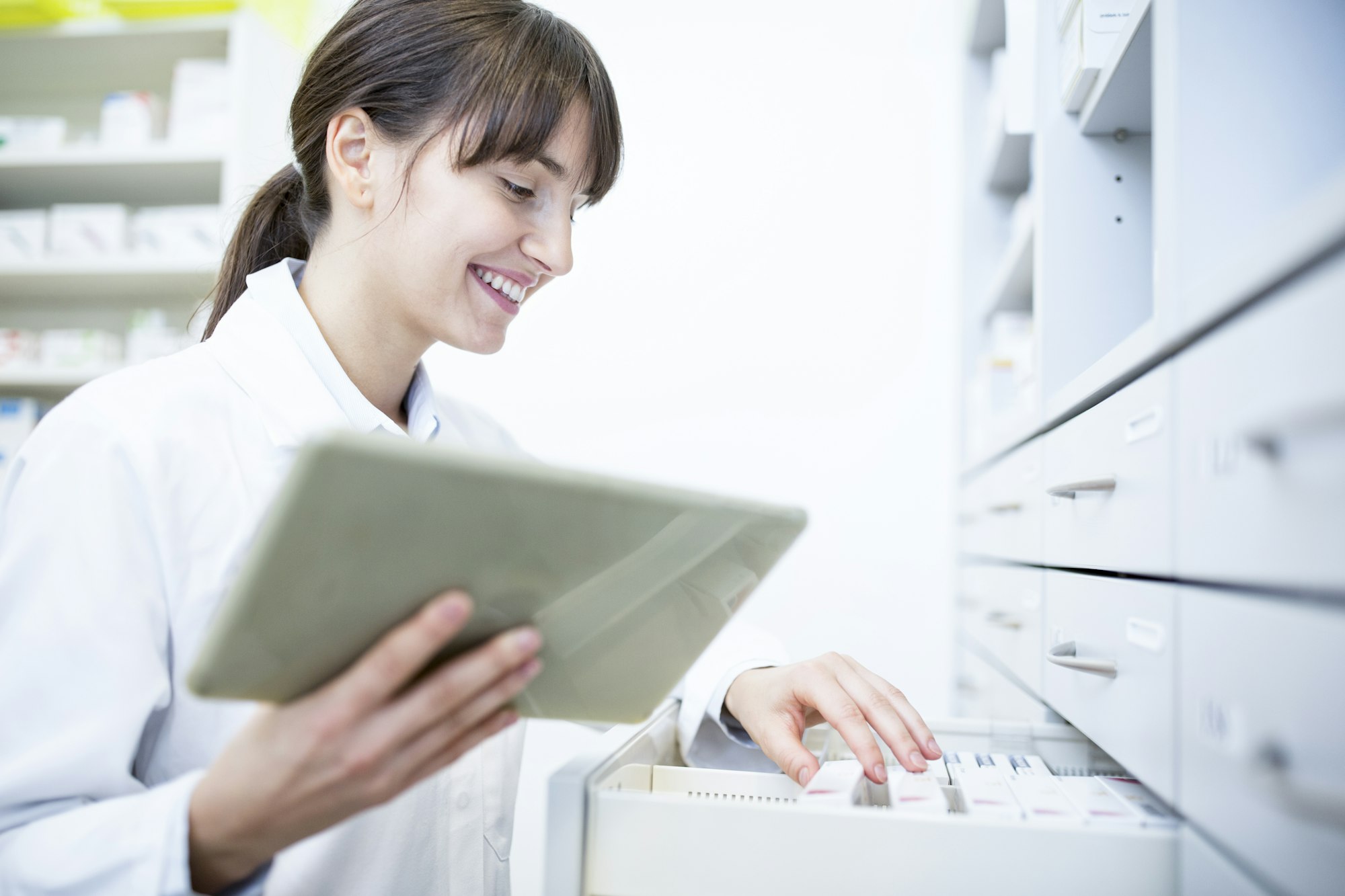 Smiling pharmacist with tablet at cabinet in pharmacy